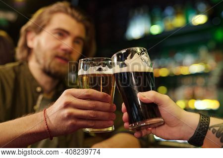 Hands of two friendly guys clinking with glasses of beer in front of camera while one of them pronouncing toast against bar with alcoholic drinks