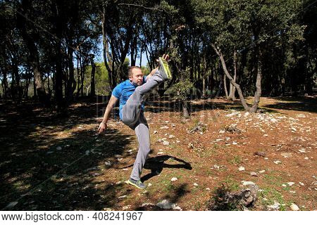 Muscular Man Doing Workout Is Warming Up By Rising Leg Up In The Air Exercise In Forest. Handsome Sp