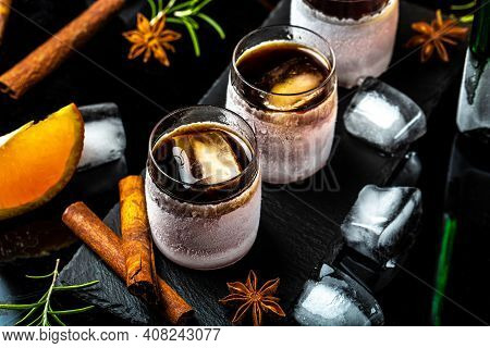 Frozen Herb Liqueur In Small Glass. Shot Glass Of Herb Liqueur With Cinnamon And Oranges On A Dark B
