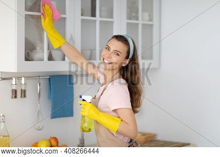 Positive Female Housekeeper Tidying Modern Apartment, Wiping Kitchen Cabinat With Detergent And Rag.