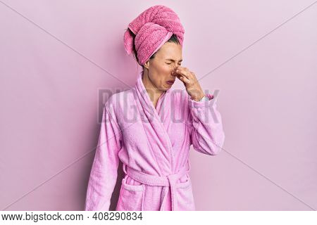 Young blonde woman wearing shower towel cap and bathrobe smelling something stinky and disgusting, intolerable smell, holding breath with fingers on nose. bad smell 