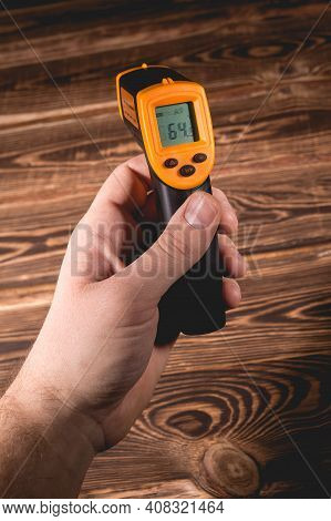 A Man Holds A Yellow-black Pyrometer On A Wooden Background. A Device For Non-contact Temperature Me