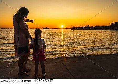 Mother Showing Her Daughter A Beautiful Sunset At The Beach While Holding Hands. Silhouette Of A Mar