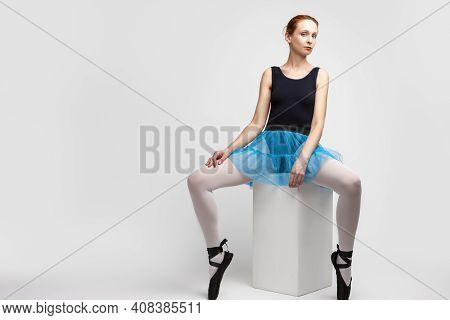 Portrait Of Tranquil Caucasian Female Ballet Dancer In Body Suit And Blue Skirt Sitting On Cube  Aga