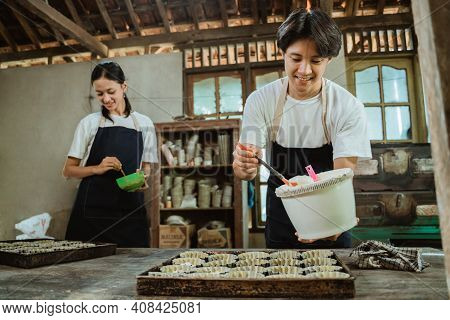 Smiling Asian Man Wearing An Apron While Pouring Dough On A Cake Pan On The Table