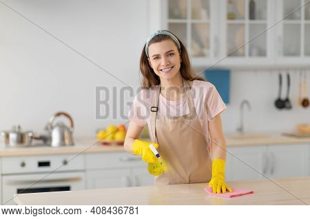 Happy Young Housewife In Rubber Gloves And Apron Cleaning Table At Kitchen, Empty Space. Positive Mi