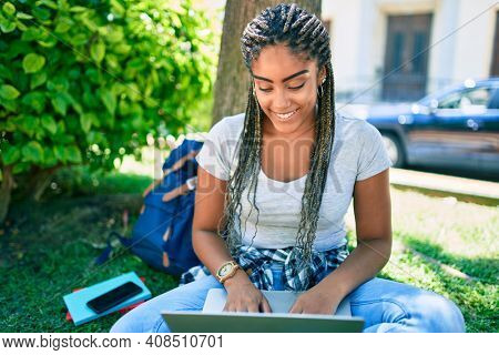 Young african american student woman smiling happy using computer laptop sitting on the grass at the university campus