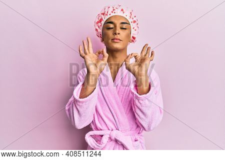 Hispanic man wearing make up wearing shower towel cap and bathrobe relaxed and smiling with eyes closed doing meditation gesture with fingers. yoga concept. 