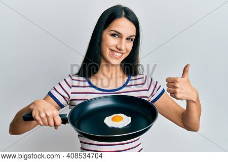 Young hispanic woman holding billet with fried egg smiling happy and positive, thumb up doing excellent and approval sign 