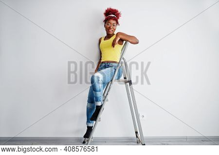 African American Woman In Yellow Singlet And Eyglasses Against White Wall Stand Om Iron Ladder.