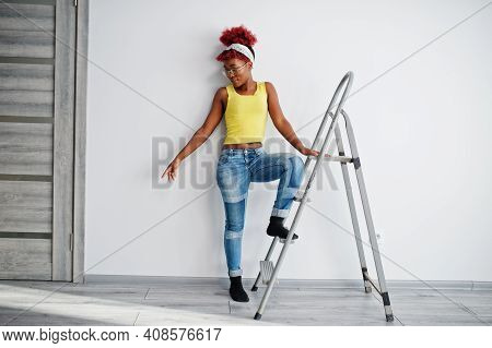 African American Woman In Yellow Singlet And Eyglasses Against White Wall Stand Om Iron Ladder.