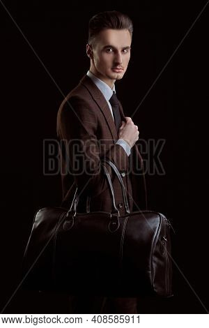 Portrait of a handsome man traveler in elegant classic suit and tie posing with leather bag on a black background. Business style. Men's fashion, accessories. Journeys.