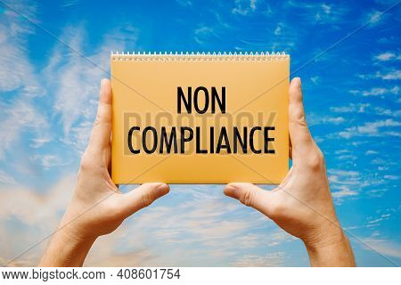 Man Holding A Card With Text Non Compliance On The Background Of A Blue Sky.