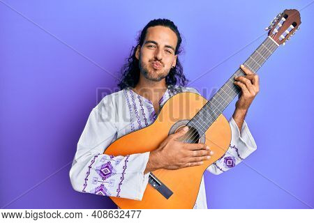Young handsome man with long hair playing classical guitar puffing cheeks with funny face. mouth inflated with air, catching air. 