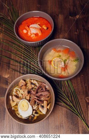 Set Of Serving Asian Soups On Gray Background Top View. Tom Yum Soup, Eel Fish Soup And Ramen Noodle