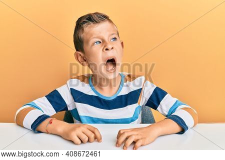 Adorable caucasian kid wearing casual clothes sitting on the table angry and mad screaming frustrated and furious, shouting with anger. rage and aggressive concept. 