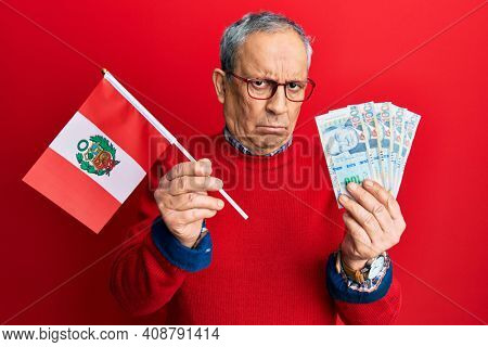 Handsome senior man with grey hair holding peru flag and peruvian sol banknotes skeptic and nervous, frowning upset because of problem. negative person. 