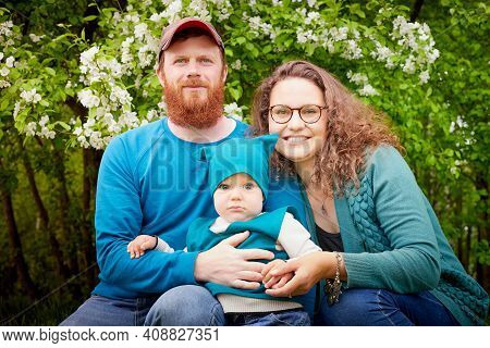 Young Family With Kid Having Picnic Outdoors Near Apple Tree With White Flowers. Parents With One Ch
