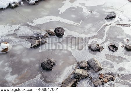 Close Up Picture Of Frozen Water With Stones In The River. Bubbles In The Icy Water. Natural Texture