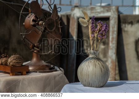 Dried Flowers In Ceramic Vase On White Textured Table Cloth And Vintage Fan With Old Cement Wall. Ho