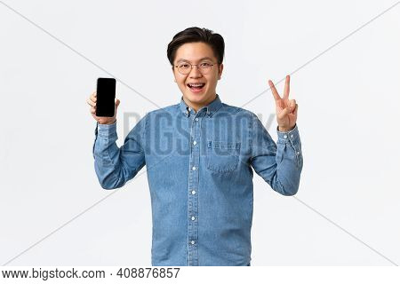 Smiling Joyful Asian Male Freelancer, Student In Glasses And Braces, Showing Smartphone Screen And P