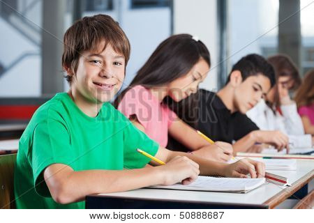 Portrait of happy teenage boy sitting at desk with friends writing in classroom