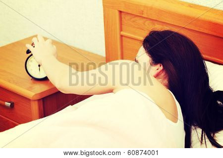 Back view closeup of a young beautiful woman reching out her hand to turn off a black alarm clock.