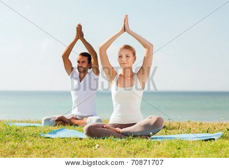 fitness, sport, friendship and lifestyle concept - smiling couple making yoga exercises sitting on mats outdoors