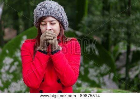 Young woman shivering with cold and blowing hot air to the hands on a forest wearing a red overcoat, a beanie and gloves during winter 