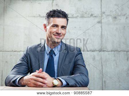 Smiling businessman sitting at the table and looking at camera over conrete wall