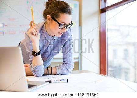 Happy young woman in glasses standing near the window in office and working with blueprint 