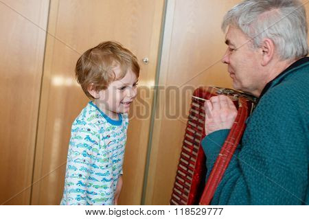Happy blond little kid boy and his grandfather playing together with accordion. Senior man teaching his grandson cute toddler to play with music instrument at home.