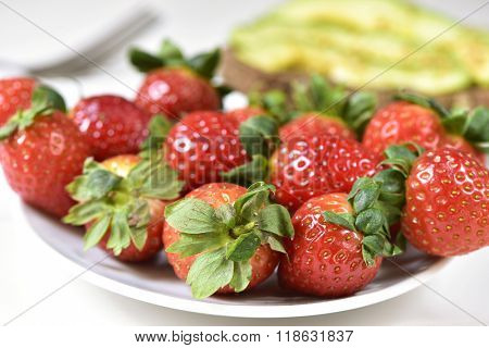 closeup of a white ceramic plate full of appetizing strawberries and an avocado toast in the background, on a set table