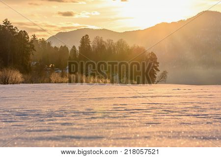 A picturesque winter landscape with trees mountains and fog over a frozen snowy lake in the rays of the sun at sunrise