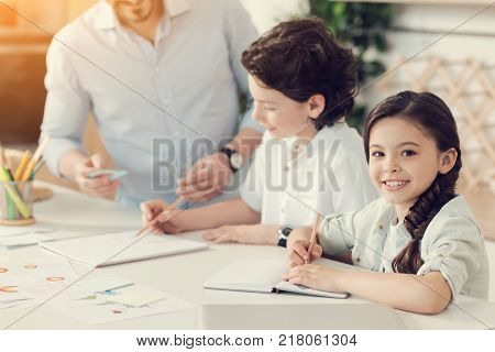 Diligent pupil. Cute positive intelligent girl sitting at the table and taking notes while doing her hometask