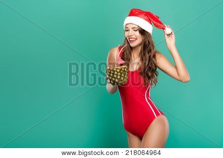beautiful woman in red bikini and christmas hat drinks summer cocktail in pineapple. christmas holiday