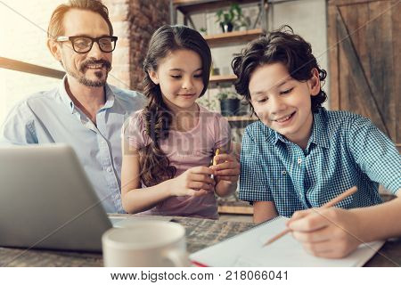 Interesting studying. Nice positive delighted children sitting at the table with their father and taking notes while doing their homework together