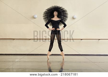 Joyful ballerina with a black tutu on her head stands on the toes in the dance hall. She wears a dark dance wear and light pointe shoes. Her hands are on the hips. Girl reflected in the glossy floor.