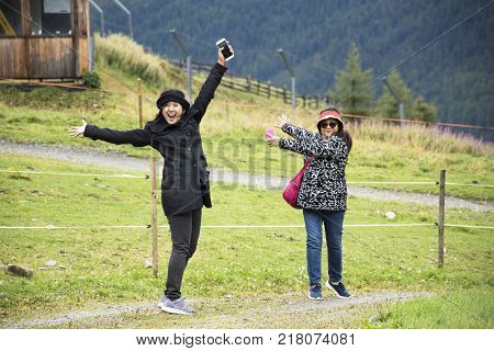 Traveler thai women mother and daughter playful travel and posing for take photo and holding mobile phone on grass field at otztal alps mountain in Schnals city in Bolzano Austria