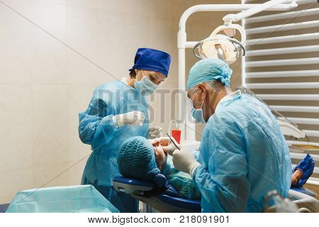 Male Old Professional Dentist Surgeon In Uniform And Female Assistant Helps To Performing Operation