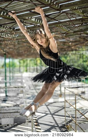 Cute ballerina hangs on her hands on the awning and reclines on the metal handrail with her feet on the concrete pier. She wears a black tutu with a leotard and looks into the camera. Outdoors.