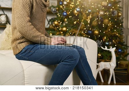 unrecognizable model using laptop at home during christmas. Buying online new year gifts. Christmas tree blur on background. Cozy apartmen decorated lights and deer
