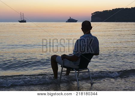 silhouette photo. a young man sits on the seashore and enjoys the sunrise. beautiful view of the bay. yacht in the background