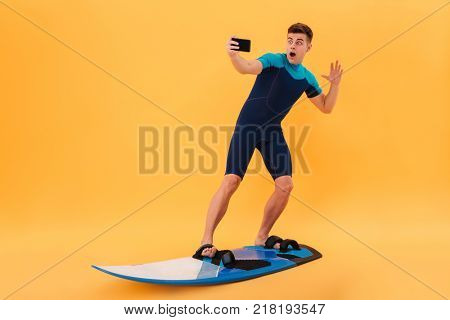 Full length photo of amased surfer guy taking selfie on smartphone while surfing on surfboard, isolated over yellow background