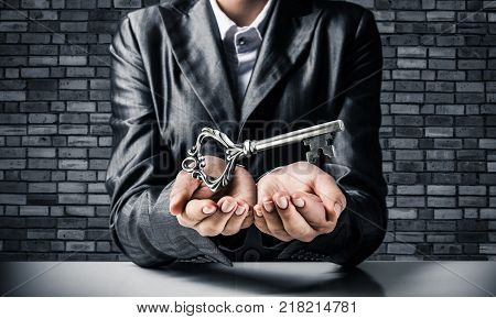 Cropped image of businessman in suit keeping big key in hands with dark brick wall on background.