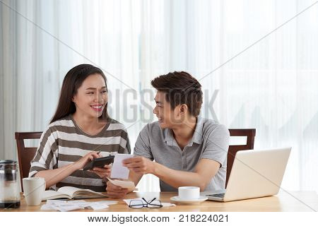 Cheerful pretty woman and her handsome husband managing family budget while sitting at wooden table, interior of living room on background