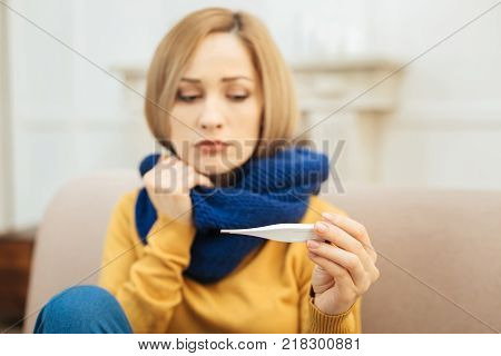 Having flu. Pretty upset young blond woman feeling down in the mouth and holding a thermometer and having a blue warm scarf on while sitting on the couch