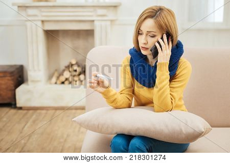 Chills. Sad young blond woman feeling bad and holding a thermometer and having a blue warm scarf on while sitting on the couch and talking on the phone