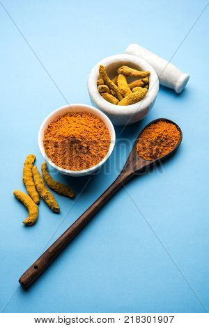 Turmeric powder in ceramic bowl with raw dried turmeric over plain background