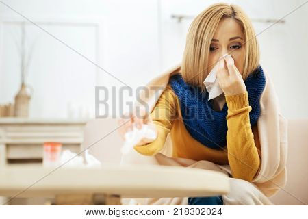 Blocked nose. Ill young blond woman blowing her nose while having a blanket on her shoulders while sitting on the couch and putting used napkins on the table in front of her